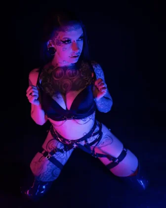 Saint Hell kniet in einem schwarzen Latex-Outfit auf dem Boden, umgeben von dramatischem Pink-Blau-Licht. Ihre Tattoos und das selbstbewusste Auftreten unterstreichen ihren dominanten Stil.
