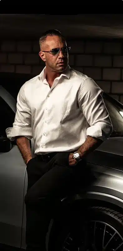 Chris Gun mit weißem Hemd und Sonnenbrille an einem grauen Auto.