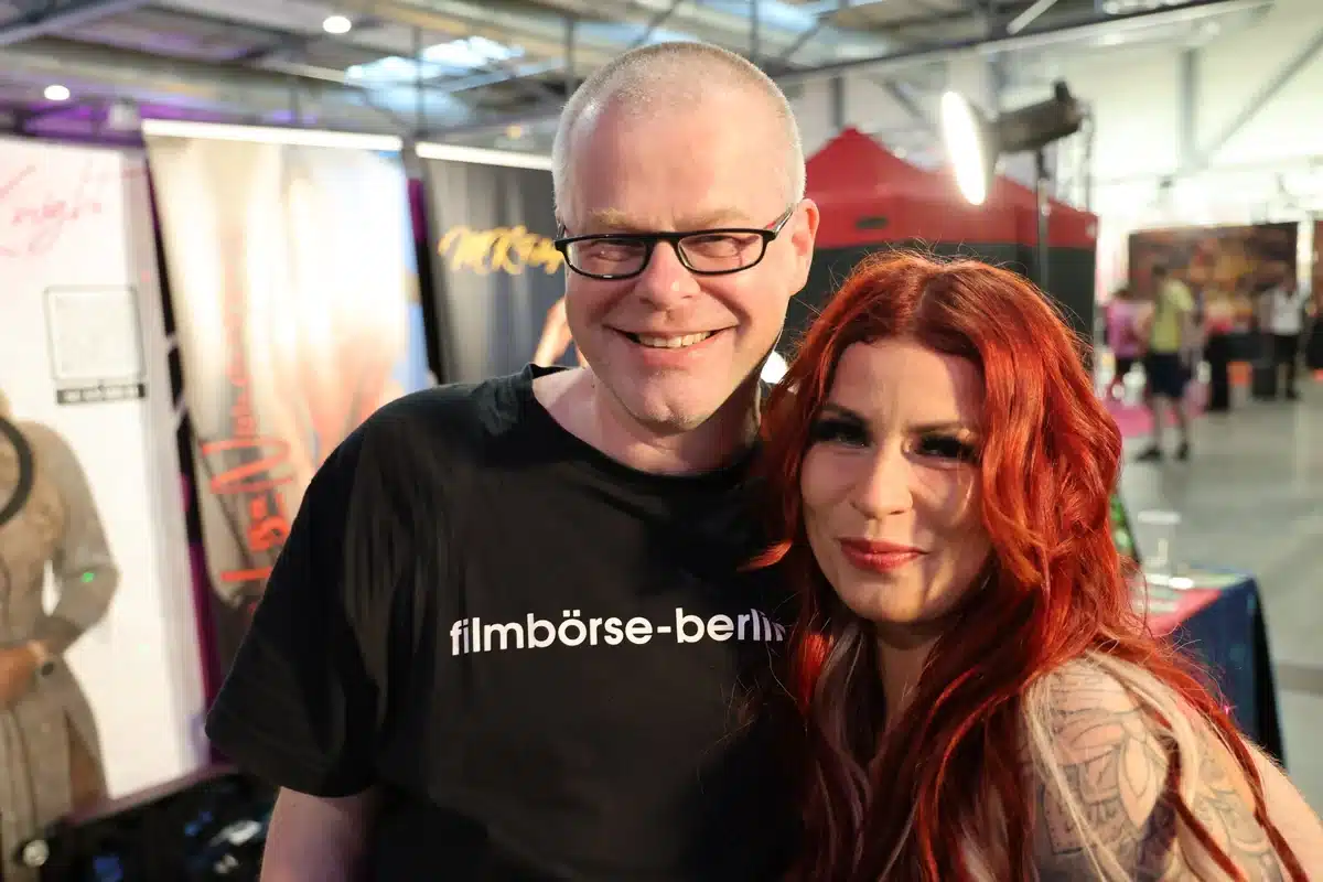 Chrissi69 mit roten Haaren und Andre Zachau im schwarzen T-Shirt lächeln gemeinsam in einer Messehalle