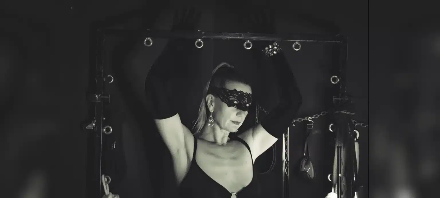 Lady Dawina Moon mit erhobenen Armen an einem schwarzen Gestell, Spitzenmaske und lange Handschuhe