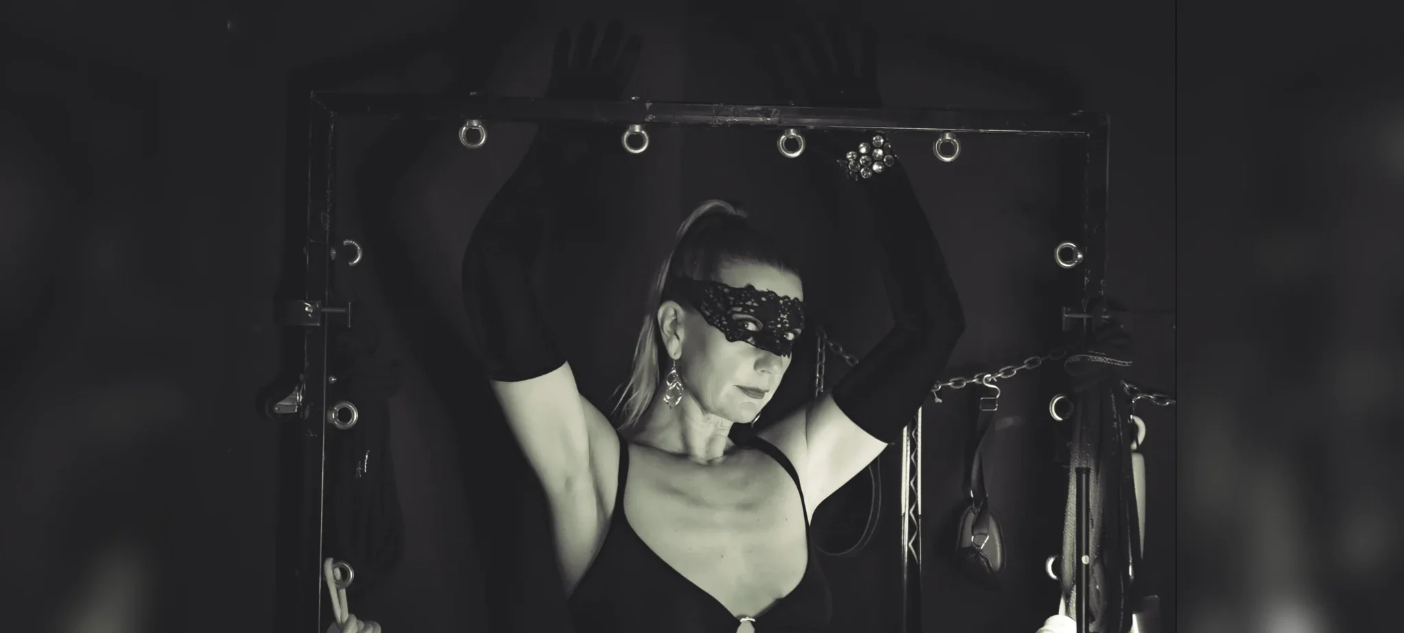 Lady Dawina Moon mit erhobenen Armen an einem schwarzen Gestell, Spitzenmaske und lange Handschuhe