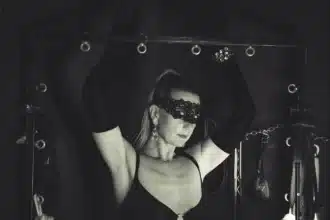 Lady Dawina Moon mit erhobenen Armen an einem schwarzen Gestell, Spitzenmaske und lange Handschuhe