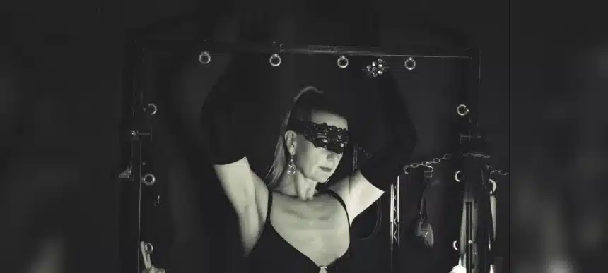 Lady Dawina Moon mit erhobenen Armen an einem schwarzen Gestell, Spitzenmaske und lange Handschuhe