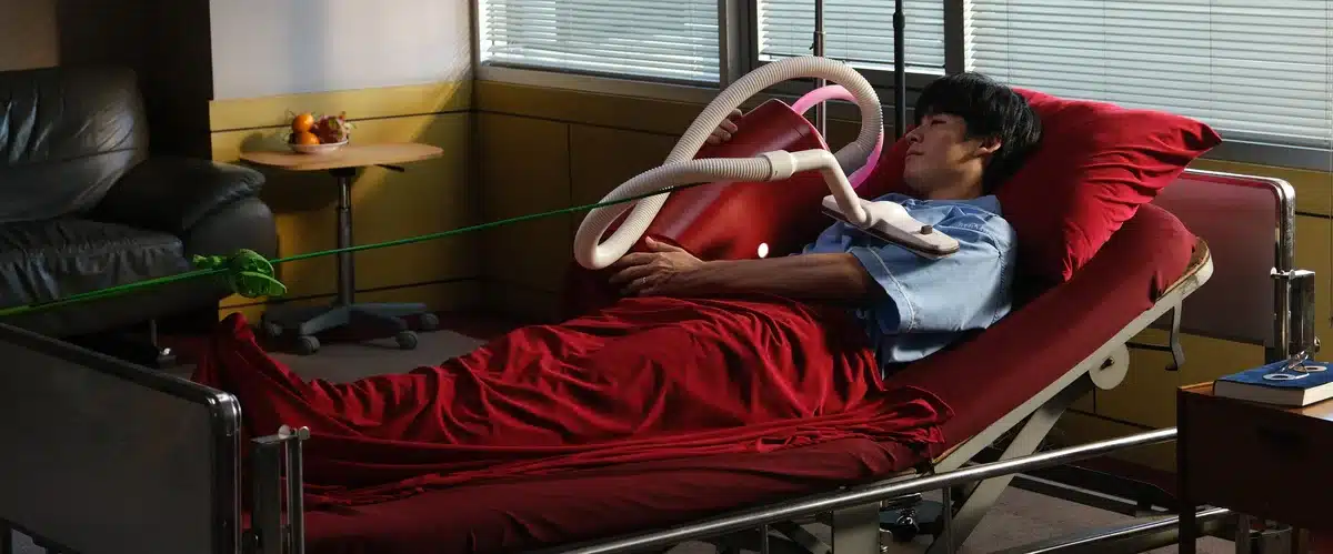 Szene aus „A Useful Ghost“: ein Mann im Krankenbett hält einen roten Staubsauger im Arm